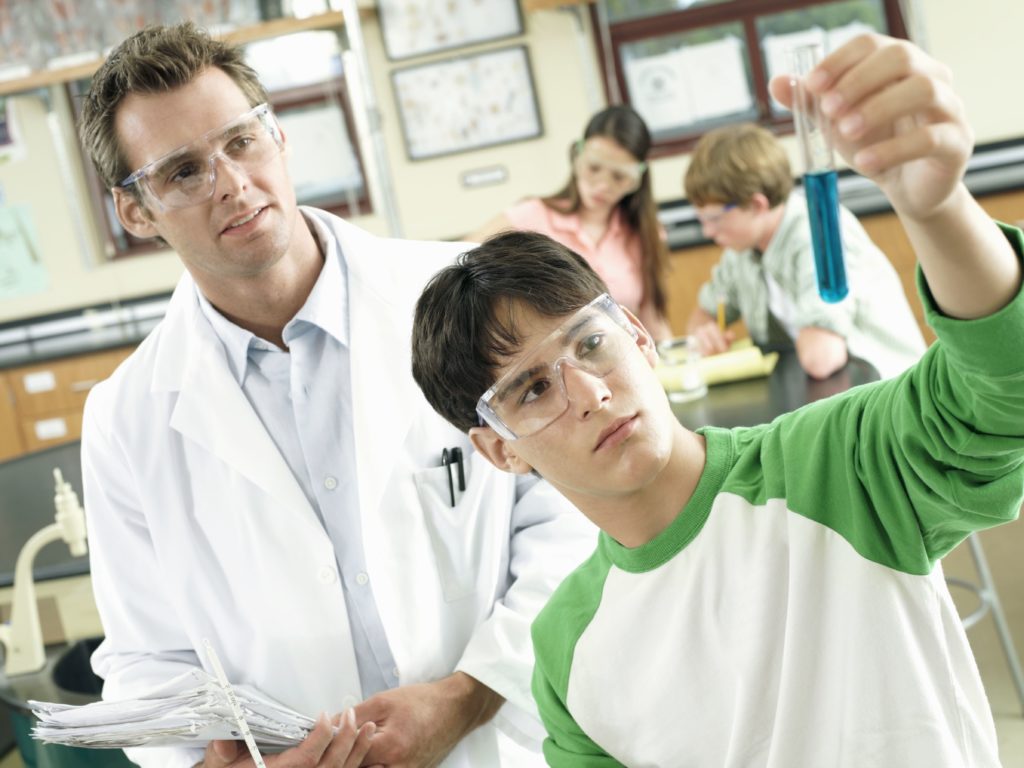 Teacher watching teenage boy (15-17) doing science experiment at desk in classroom (tilt)